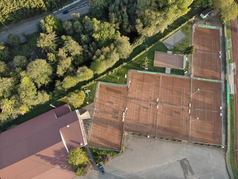 Luftaufnahme von mehreren Tennisplätzen mit rotem Sand. Neben den Tennisplätzen steht ein Gebäude, dahinter stehen Bäume.