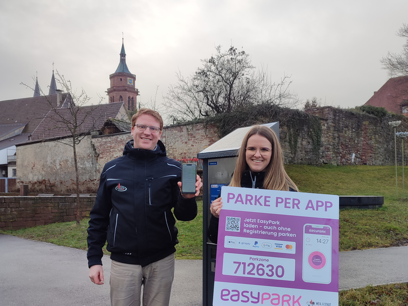 Zwei Verwaltungsmitarbeiter machen mit einem Smartphone und einem Schild in der Hand WErbung für die Park-App.