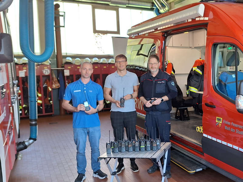 Bürgermeister Walter, Stadtbrandmeister Bäuerle und ein Mitarbeiter präsentieren vor einem Feuerwehrauto die neuen Funkgeräte.