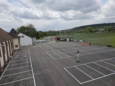 Ein Parkplatz mit weißen Markierungen. Am Rand steht die Turn- und Festhalle, auf der anderen Seite ein Fußballplatz.