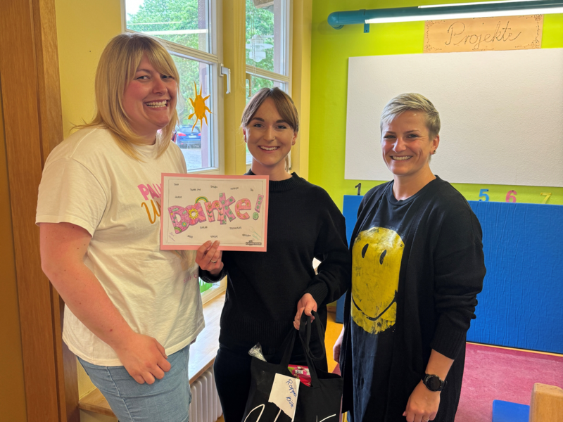 Drei Frauen stehen in einem bunten Raum eines Kindergartens. Eine Frau hält eine Karte hoch, auf der DANKE steht.