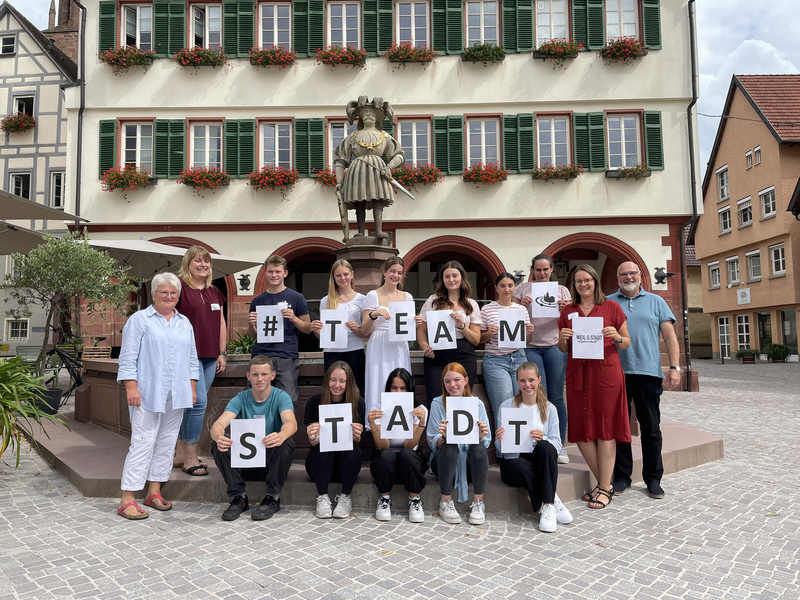 Die Nachwuchskräfte und Verwaltungsmitarbeiter stehen vor dem Rathaus. Die Schilder in ihren Händen bilden den Schriftzug TEAM STADT.