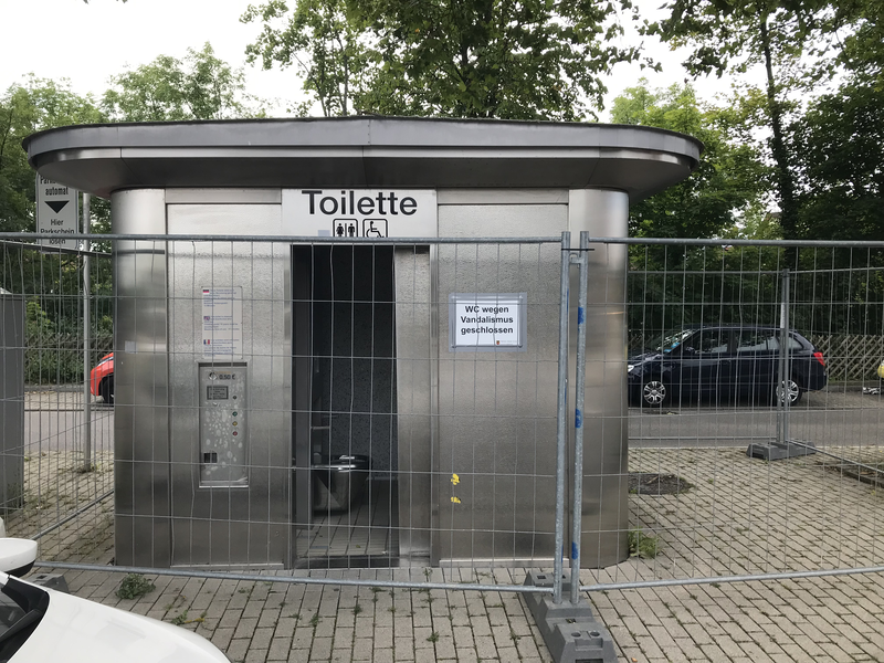 Öffentliche Toilette mit geöffneter Tür, umzäunt und mit einem Schild "WC wegen Vandalismus geschlossen".