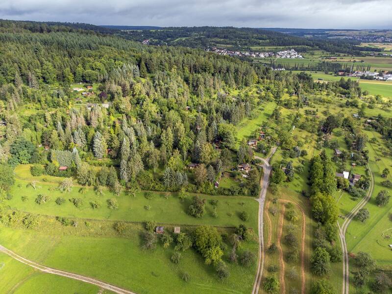 Eine Luftaufnahme zeigt eine weitläufige Landschaft mit dicht bewaldeten Flächen, vereinzelten Wiesen, kleinen Wegen und Gartengrundstücken.