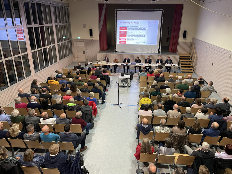 Viele Menschen sitzen auf Stühlen in einer Halle. Vertreter der Stadtverwaltung sitzen vorne an Tischen und zeigen eine Präsentation.