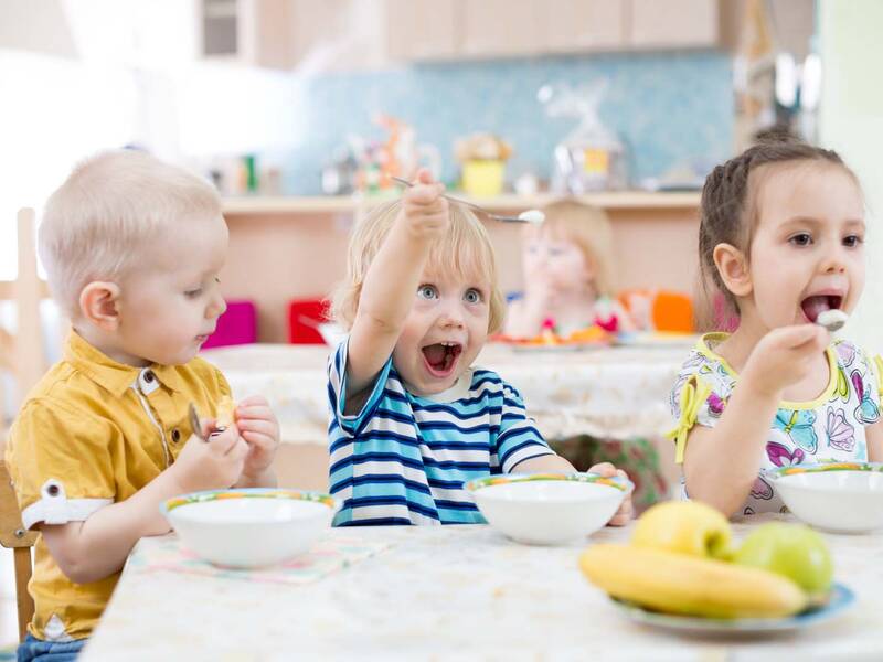 Drei Kinder sitzen an einem Essenstisch. Vor den Kindern stehen Schüsseln auf dem TIsch sowie ein Teller mit Obst. Das rechte Kind hält einen Löffel in der Hand und schaut in die Schüssel neben sich. Das Kind in der Mitte hebt sein Löffel in die Luft. Das rechte Kind führt den Löffel in sein Mund rein. Im Hintergrund sitzt ein weiteres Kind an einem Tisch. Hinter diesem Kind ist eine Küche.