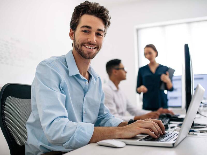 Ein Mann arbeitet an seinem Laptop. Im Hintergrund ein Büro mit weiteren Personen.