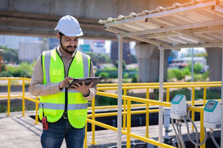 Ein Mann in Warnweste steht mit einem Tablet in der Hand auf einer Baustelle.